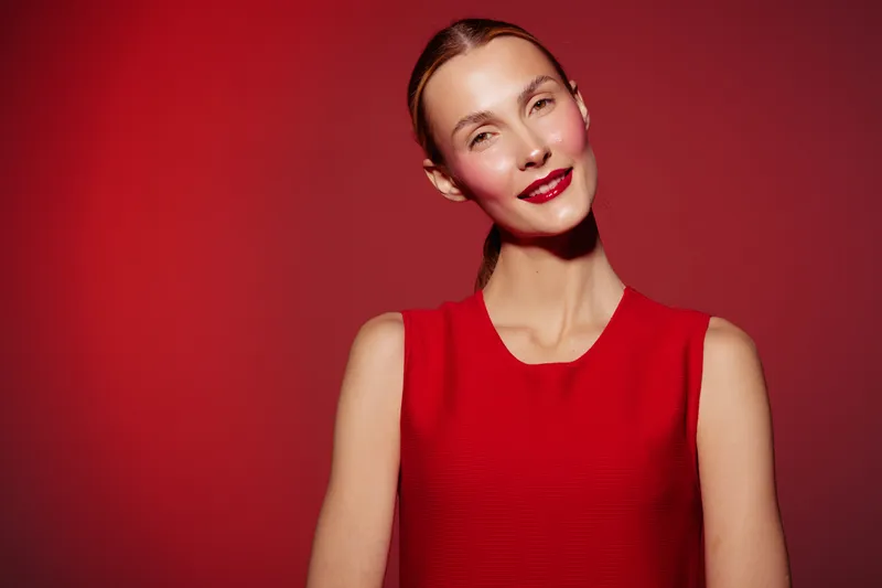 Smiling woman in red sleeveless dress poses against red background. Studio portrait with soft lighting highlights natural beauty and confident expression. Concept of fashion and elegance.