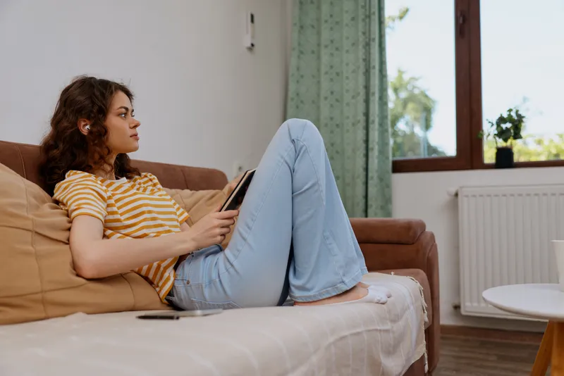 Young woman with curly hair relaxing on a sofa using a tablet at home. Bright daylight streams through a window, creating a calm, cozy living room scene focused on comfort.
