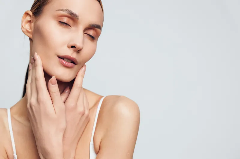 Young woman with closed eyes gently touches her face with fingers showing natural skin beauty and calm expression isolated on plain light background in studio.