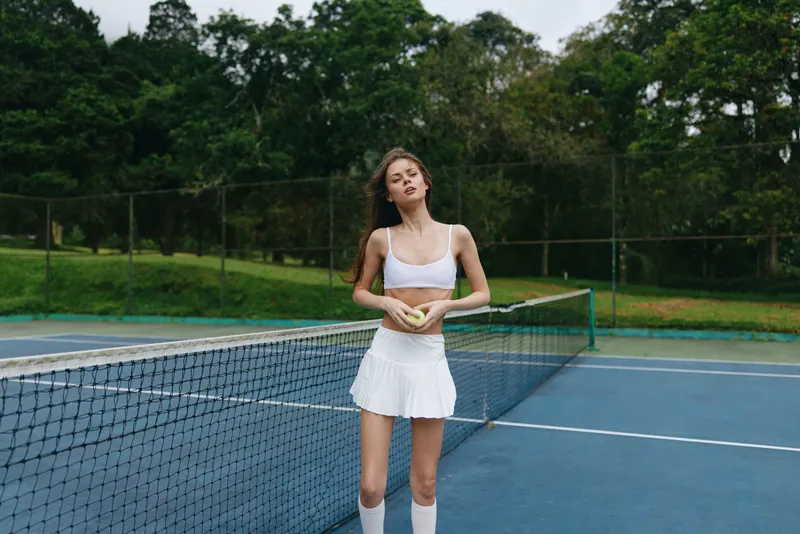 Young female tennis player in a sports outfit confidently posing on the court, showcasing athleticism and determination, surrounded by nature and greenery