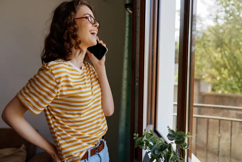 A woman in a striped tshirt stands by a sunlit window, talking on a phone in a cozy home setting. Casual lifestyle moment, everyday communication, warm, relaxed mood.