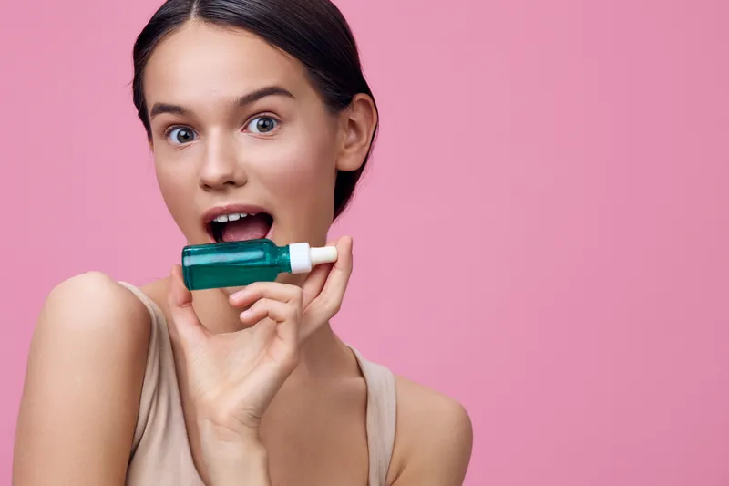 Young woman with healthy skin holding a green skincare bottle, smiling joyfully on a pink background, representing beauty and self care