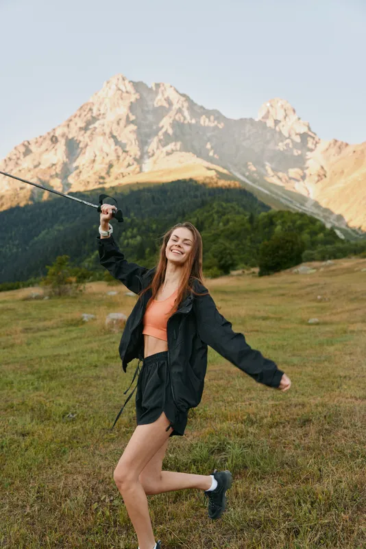 A cheerful young woman enjoys outdoor adventure in a mountain meadow. She smiles brightly, wearing a light jacket and shorts, radiating freedom, energy, and playful spirit.