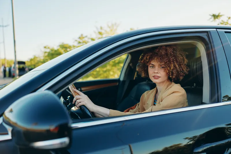 Young woman with curly hair drives a sleek car under a bright sun, enjoying a relaxed moment behind the wheel on a quiet road with warm retro tones.