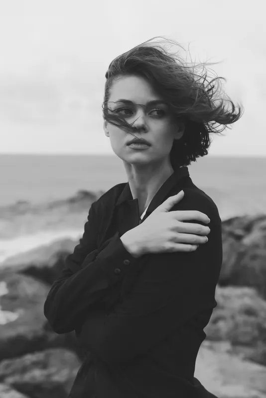 Windswept woman by the coastline in a dark coat, moody black and white scene showcasing fashion, silhouette, and coastal atmosphere for editorial storytelling