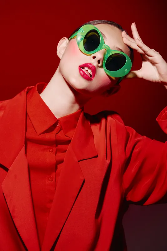 Fashionable woman wearing bright red coat and green round sunglasses poses against a red background. Bold makeup and strong style create a creative beauty and fashion concept.