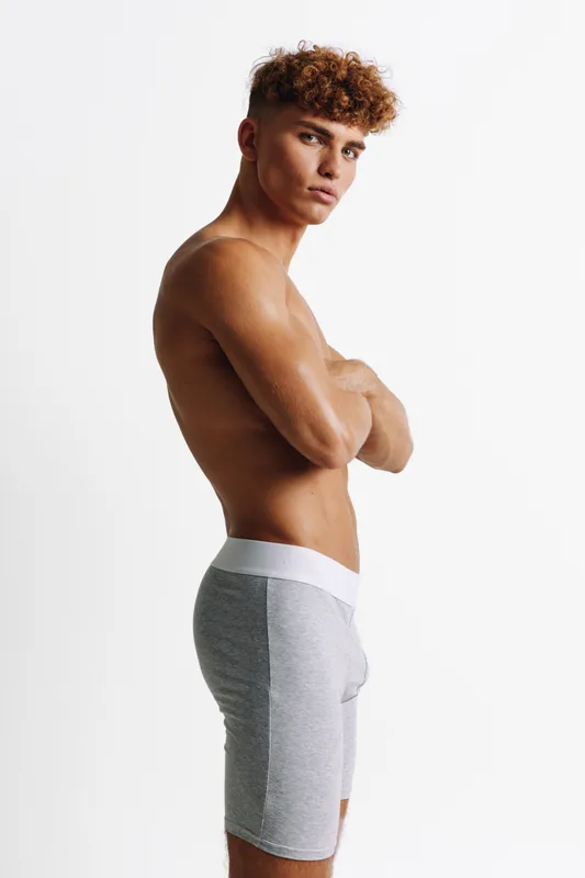 Young athletic man with curly hair posing shirtless in gray shorts against white background, showing confident expression and muscular build in studio.