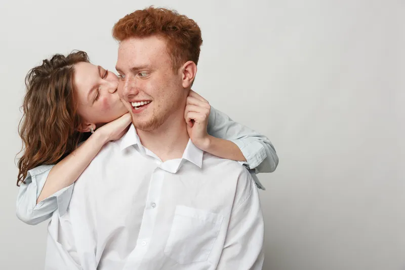 Happy young couple in casual clothes share affectionate moment, woman hugs man from behind and kisses his cheek. Concept of love, relationship, intimacy, and happiness together.
