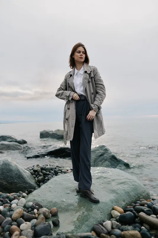 A confident woman stands on rocky shore wearing a textured coat and dark trousers. Overcast sky and calm sea create a serene and contemplative outdoor mood.