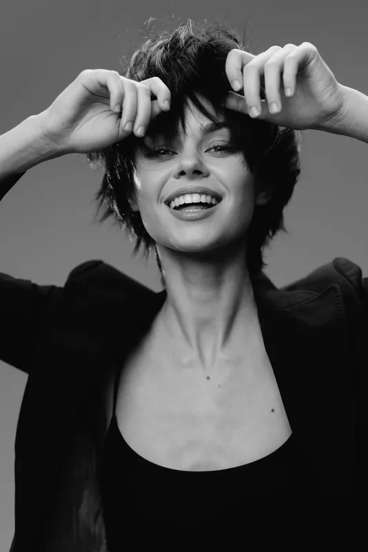 Smiling woman with short hair in black and white style, expressing joy and carefree attitude, wearing dark clothing against neutral background.