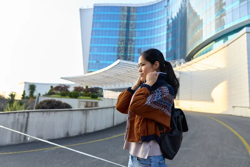 Woman with backpack on an urban outdoor walkway near glass skyscrapers adjusting hair wearing casual jacket capturing a street style moment in daylight