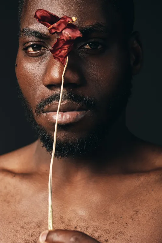 Creative visual portrait of dark skinned African man holding dried red flower in front of face with serious expression in studio. Artistic closeup with bare chest and black background.