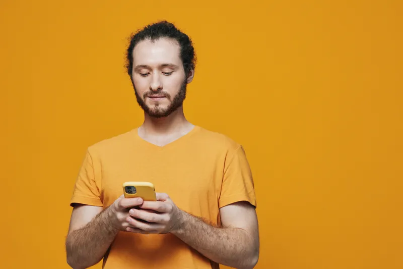 Young man using a smartphone against a vibrant yellow background, wearing a casual orange t shirt, showcasing a focused expression, ideal for technology and communication themes