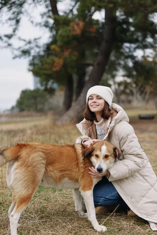 Woman in a warm coat kneels beside a friendly dog in a park, sharing gentle companionship on a cool day, autumn trees in the distance and soft natural light on a calm outdoor moment