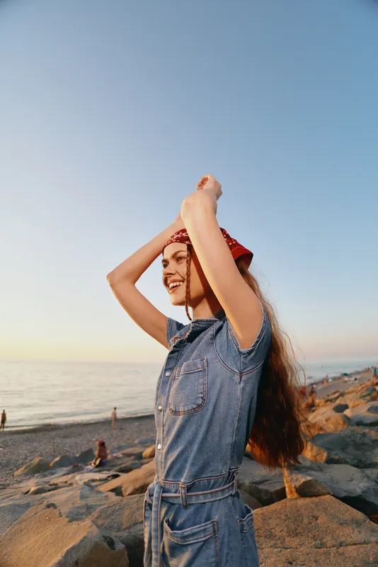 Lifestyle woman wearing modern boho-western denim street style outfit smiling outdoors near rocky beach during warm sunset with film color tones and casual carefree mood