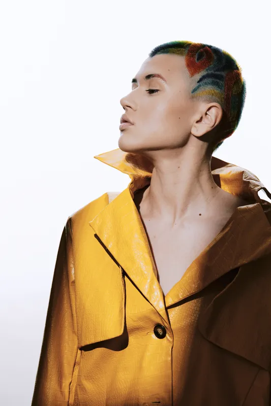 Woman with colorful short hair and floral design, wearing a yellow coat, looking sideways with a confident expression on a bright white background.