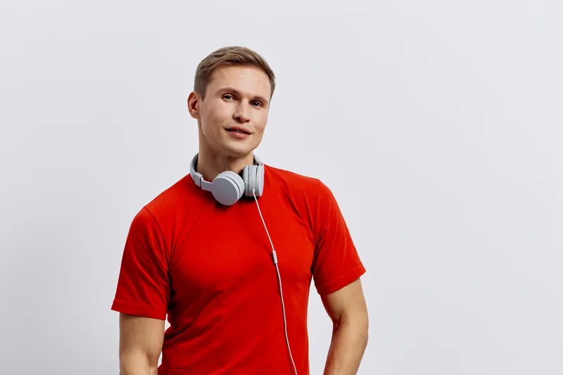 Casual young man with short blond hair smiling, wearing red t-shirt and headphones around neck, standing against plain white background. Lifestyle, relaxed, music lover, casual wear, studio shot, cont