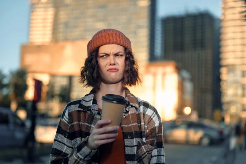 Young person holding coffee on urban street, beanie and plaid jacket, skeptical expression captured in a candid moment with authenticity, golden hour glow, mindful living, emotional storytelling.