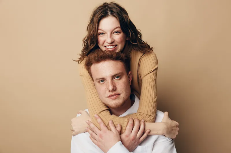 Smiling young woman with curly hair hugs serious redhead man from behind on beige background. Concept of friendship, affection, and casual lifestyle in studio portrait photography.