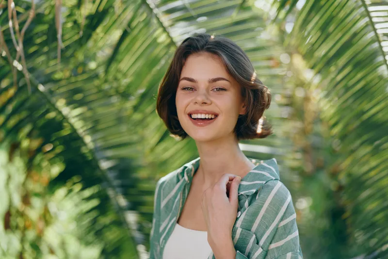 Portrait of a cheerful young woman outdoors among tropical palms, wearing a striped green shirt and white top.