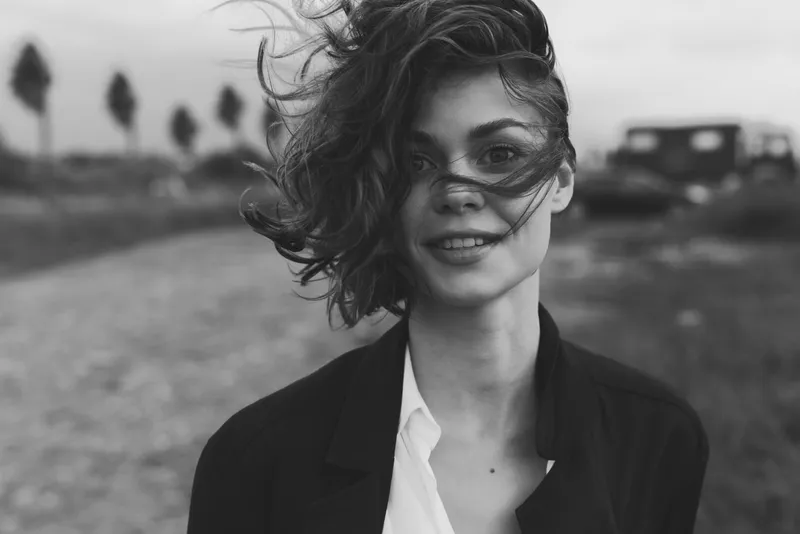 Woman smiling in a windy outdoor setting, wearing a blazer, moody monochrome scene, candid style and natural charm.