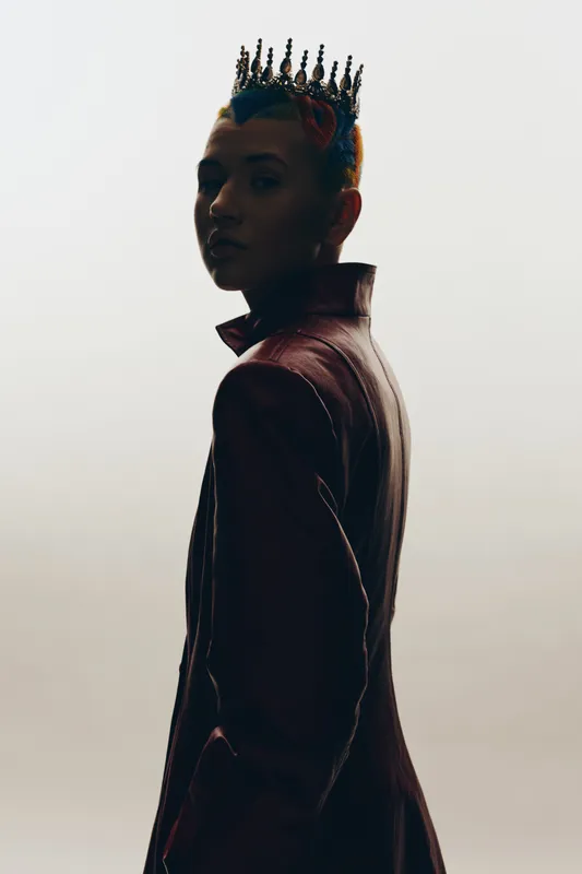 edgy woman with crown and vibrant hair styled in a leather jacket, striking a moody pose against a minimalistic gray background, artistic portrait concept.