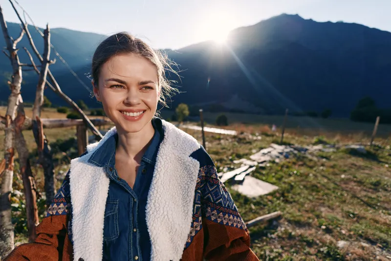 Woman smile mountains sunrise outdoors portrait wearing patterned jacket and denim shirt, happy traveler enjoying scenic nature landscape with sunburst and valley behind in morning light.