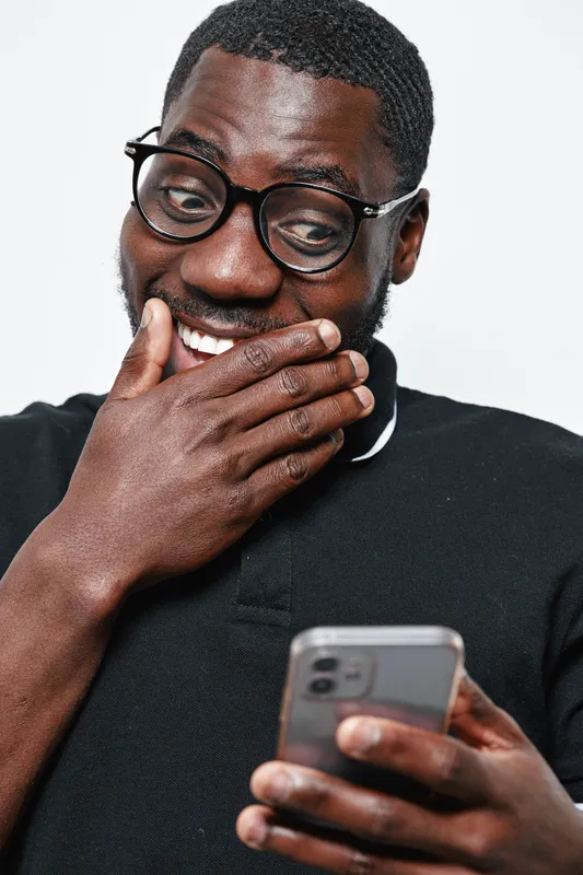 Black man with dark skin and glasses smiling and covering mouth while looking at smartphone screen in casual black shirt on white background.