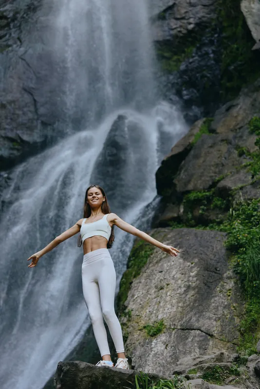 young woman standing with arms outstretched near a waterfall wearing white activewear surrounded by rocky nature, expressing freedom and peace in a serene outdoor setting