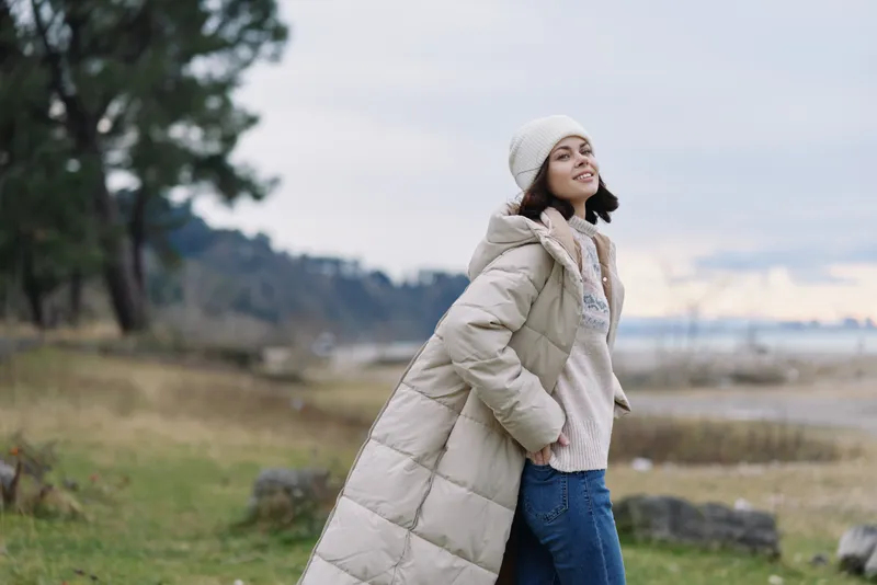 Woman outdoor coat winter fashion in countryside landscape, walking by the coast with beige puffer, jeans and beanie, calm scene with open field and soft light