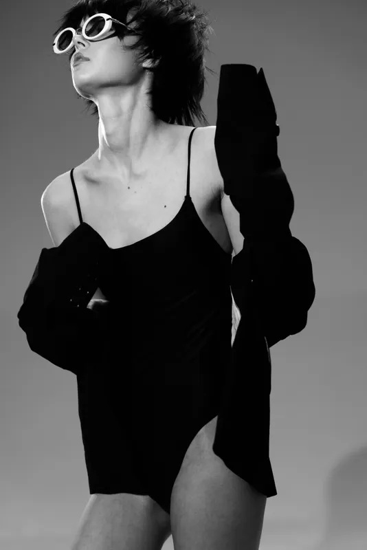 Black and white portrait of a stylish woman wearing sunglasses and a black dress with jacket, expressing confident and edgy fashion attitude.
