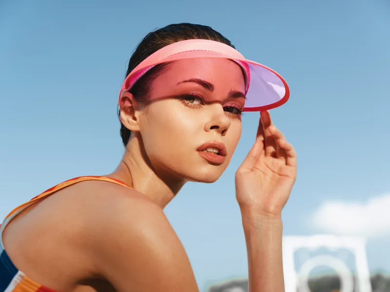 Portrait of a stylish athletic woman wearing a pink visor under bright sun, closeup perspective, confident expression, smooth skin, vibrant summer tones, and dynamic energy in an outdoor setting.
