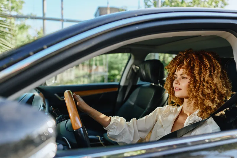 Young curly woman driving car confidently, wearing seatbelt and light blouse, with natural outdoor background and bright daylight lighting.