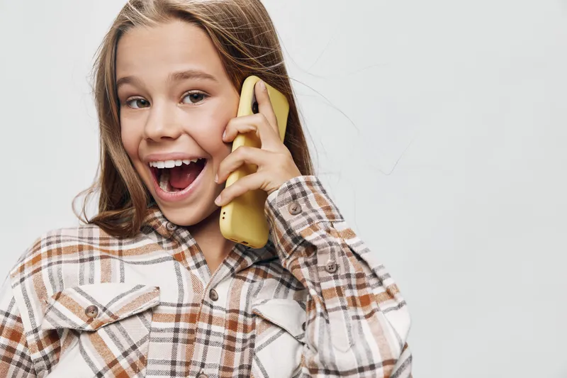 Girl smiling and talking on smartphone with excitement in casual shirt on gray background. Happy teenager with long hair holding yellow mobile phone for communication.