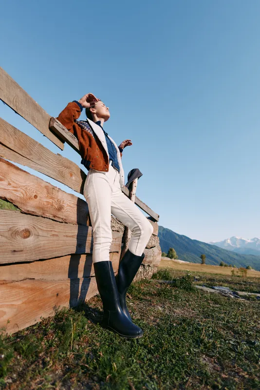 Person standing by wooden fence in rural field, wearing boots and cozy sweater, looking up at clear blue sky with distant mountains, relaxed outdoor lifestyle and country fashion.