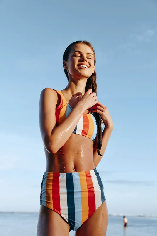 Smiling woman in a striped swimsuit on a sunny beach, standing near the ocean, eyes closed in warm sunlight, enjoying summer vacation, bright blue sky, outdoor lifestyle, summer breeze