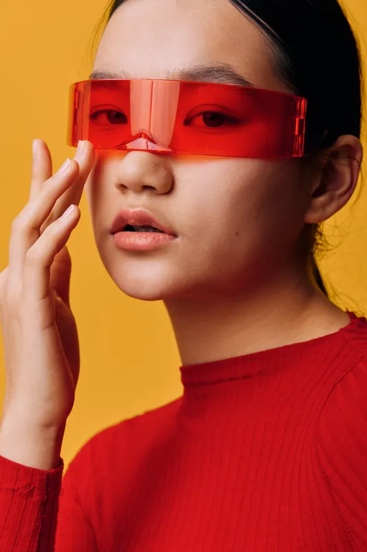 Woman wearing futuristic red glasses and a red sweater touching her face with her hand against a yellow background with sleek hair and smooth skin closeup portrait.