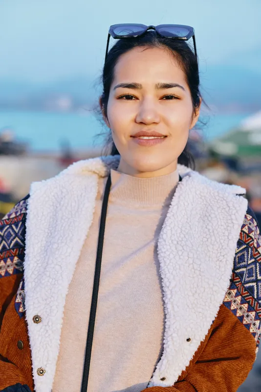 Woman outdoors by a sunny coastline wearing a patterned jacket and fluffy vest, sunglasses on head, calm smile, casual fashion, blue sea and sky backdrop, candid lifestyle shot for travel and leisure