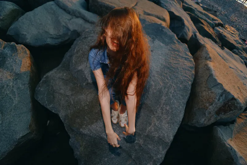 Lifestyle woman in modern boho-western denim street style with long red hair climbing rocks outdoors, captured in warm film color tones for a natural, adventurous vibe.