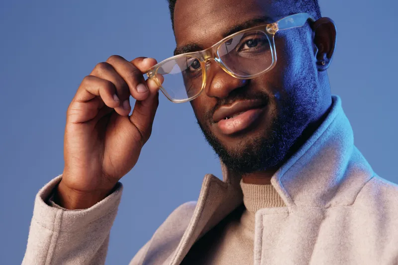 Handsome Black man wearing stylish glasses and a modern coat, smiling confidently against a blue background, showcasing contemporary fashion and elegance