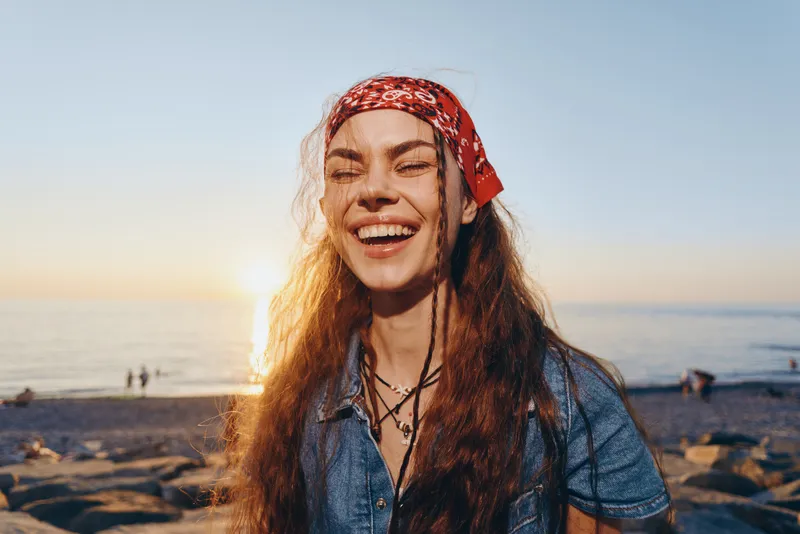 Lifestyle woman in modern boho-western denim street style smiling on sunny beach with warm film color glow and a red bandana in her hair during golden hour.