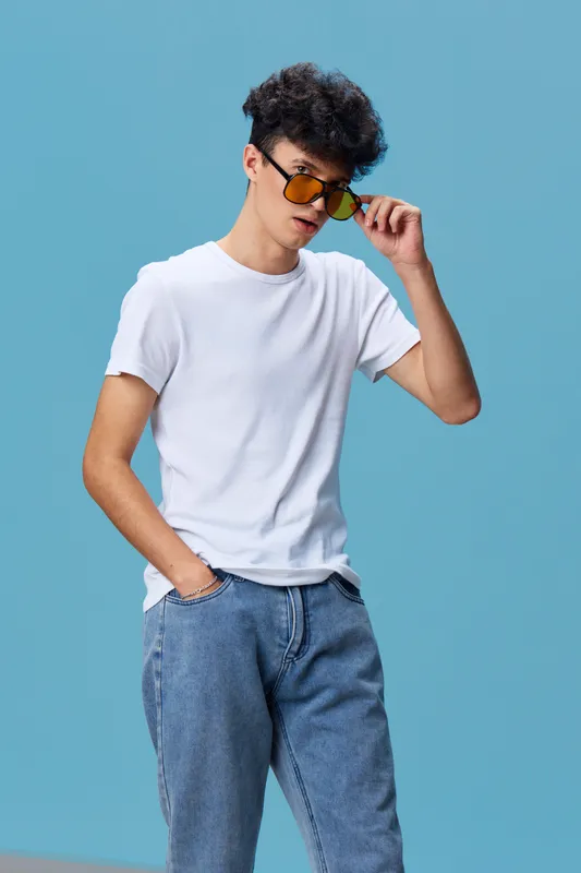 young man wearing yellow sunglasses and casual white t-shirt with blue jeans posing confidently against blue background, stylish hairstyle, modern fashion look