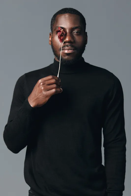 African American man with dark skin holding dried flower in front of eye in creative visual design style. Portrait with black turtleneck and neutral gray background for artistic concept.
