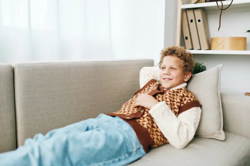 A young boy lounges on a soft sofa, wearing a patterned knit sweater, smiling as he relaxes in a cozy living room with gentle natural light.