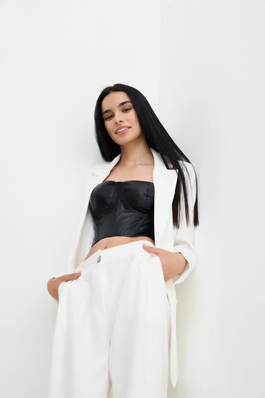 Young woman in stylish white suit and black corset posing confidently with hands in pockets against pure white background, showcasing modern fashion elegance.