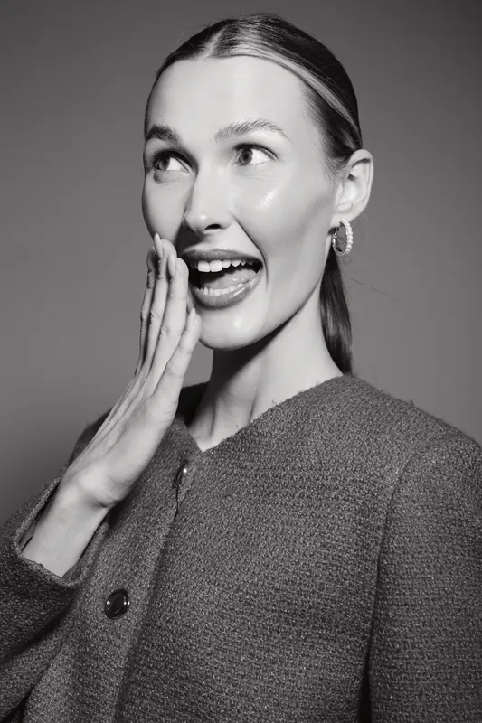 Young Caucasian woman with light skin and slicked back hair poses surprised in grey textured jacket on plain background in black and white photo.
