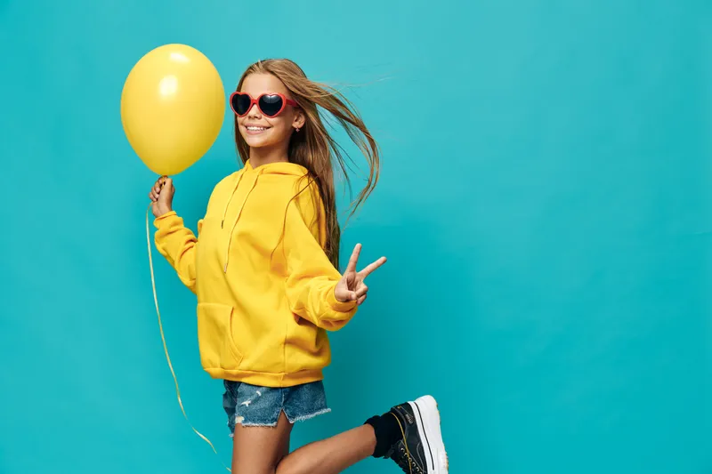 Girl in yellow hoodie holding balloon and making peace sign with hand while jumping against bright blue background. Child wearing heart-shaped sunglasses and denim shorts smiling.