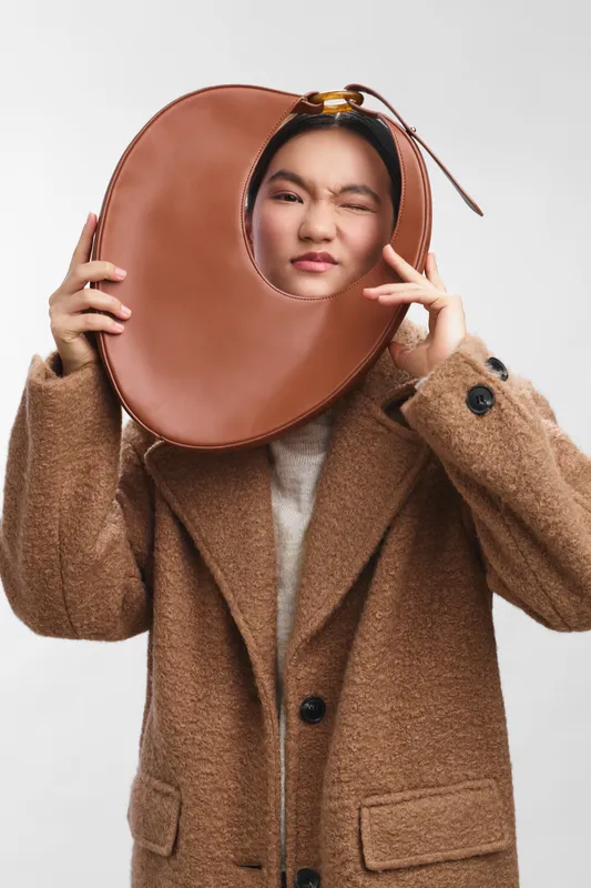 Asian woman wears a stylish brown coat with a circular leather bag around her head, posing playfully in a studio. The quirky accessory and confident expression create a bold, fashion forward portrait.