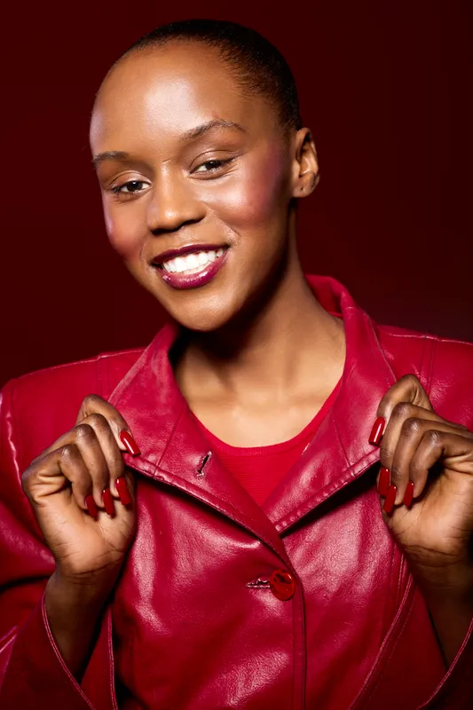 Smiling black woman with dark skin wearing a red leather jacket poses confidently on a red background. Concept of fashion, beauty, confidence, and style with vibrant colors.
