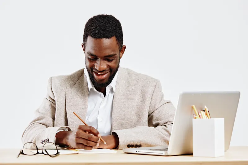 Confident young African American man working at desk using pen and laptop in modern office setting. Happy, focused expression, casual business attire, professional environment, bright minimalistic bac